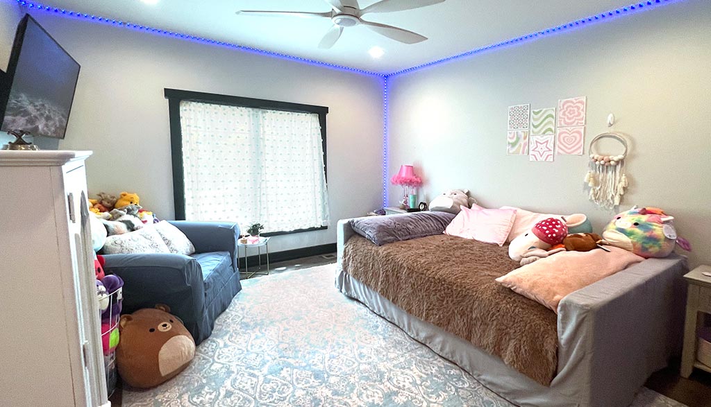 Wide angle photo of a girl's bedroom with an armchair in the corner, a bed, and blue lights framing the room